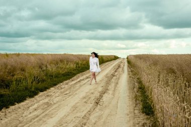 A mesmerizing woman in a white dress gracefully walks along a sun-kissed dirt road, surrounded by golden fields under a cloudy sky, creating a serene and idyllic scene of rural beauty .