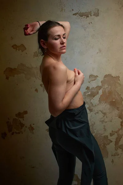 A topless woman in black pants poses confidently against a textured wall. The image showcases her beauty and strength with orange nail polish and the interplay of light and shadows.