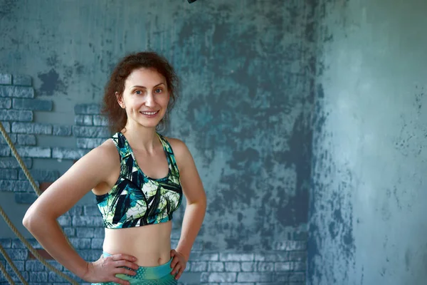A smiling brunette woman in sportswear stands confidently in a fitness studio with a textured wall and climbing rope. She is wearing a sports bra and leggings. The shot is waist up.  