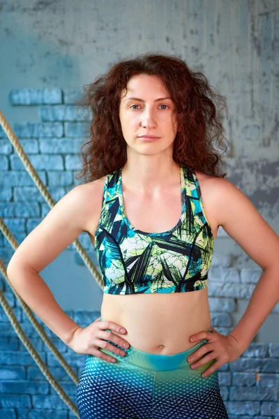 An athletic woman in sportswear stands confidently with hands on hips in front of a blue brick wall. She appears fit and healthy, possibly after a workout. 