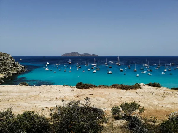 Golden sandy bay with vivid turquoise water and many small boats anchored offshore.