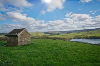 Vibrant green fields stretch towards a peaceful river, with a rustic stone structure nestled on a hilltop. The sky features fluffy clouds illuminated by sunlight, creating a tranquil atmosphere.