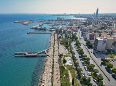 The vibrant waterfront promenade features lush greenery and a busy road alongside the sparkling sea, with a bustling harbor visible and a modern skyline in the background.
