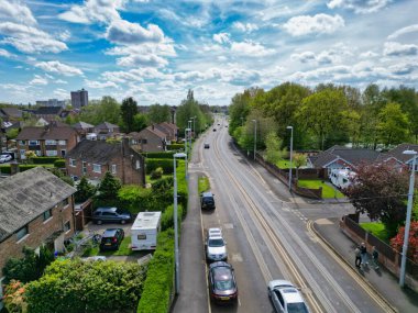 Vehicles travel along a residential street, featuring lush green trees and homes. The bright clouds add a serene feel to the urban landscape. People walk along the sidewalk, enjoying the day.