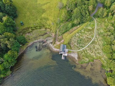 A tranquil lakeside scene features a rustic dock at the waters edge. Lush greenery surrounds the area, accompanied by a charming path leading into the woods.