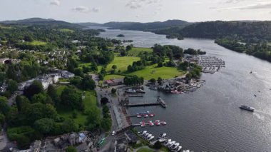 A breathtaking panorama captures a tranquil lakeside harbor surrounded by vibrant green hills. Boats dot the water while lush trees frame the scene on a bright day.