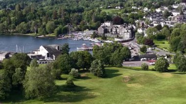 A picturesque setting showcases a charming lakeside town bustling with people enjoying various activities. Green hills surround the area, enhancing the serene atmosphere.