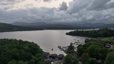 As dusk settles in the Lake District, a tranquil lake view unfolds, showcasing anchored boats and lush greenery. The dramatic sky adds to the peaceful atmosphere, inviting relaxation.