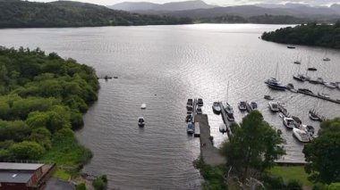 A peaceful lakeside view features several boats moored at a dock. Lush greenery surrounds the area, with rolling hills visible in the background under a cloudy sky.