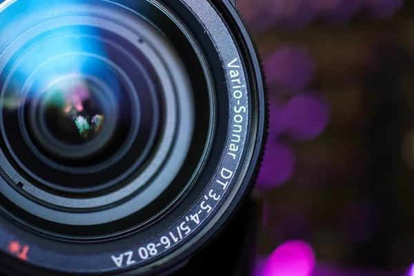A camera lens is prominently displayed, highlighting its detailed construction. The colorful, blurred background adds depth, enhancing the focus on the lens itself.