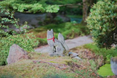 A stone fox statue sits gracefully in a serene Japanese garden, surrounded by lush greenery and a well-maintained pathway, capturing a moment of peaceful contemplation.