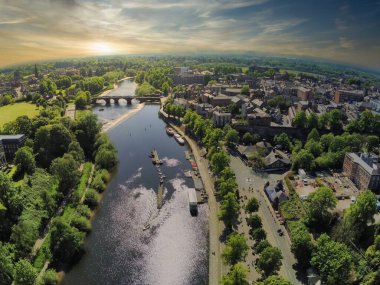 Overlooking the River Dee at sunset, lush trees line the banks while historic buildings can be seen in Chester. The tranquil waters reflect the warm colors of the evening sky.