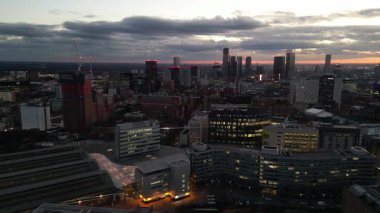 A breathtaking view of the Manchester skyline as dusk falls. City lights begin to twinkle while clouds drift across the sky. The setting sun casts a warm glow over the urban landscape.