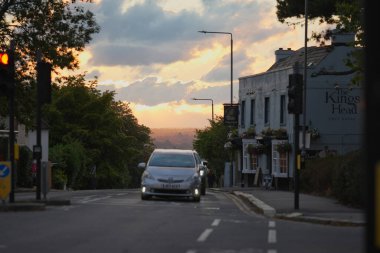 A silver car drives along a peaceful road as the sun sets in the background. The warm colors of the sky create a beautiful atmosphere near a local pub.