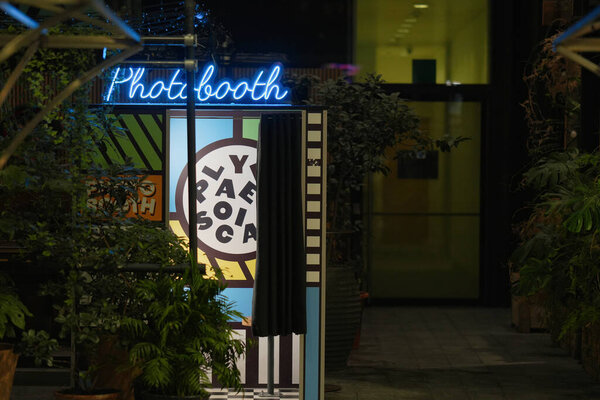Colorful photo booth stands in a vibrant location, surrounded by lush plants. The inviting neon sign enhances the lively atmosphere, perfect for capturing memories.