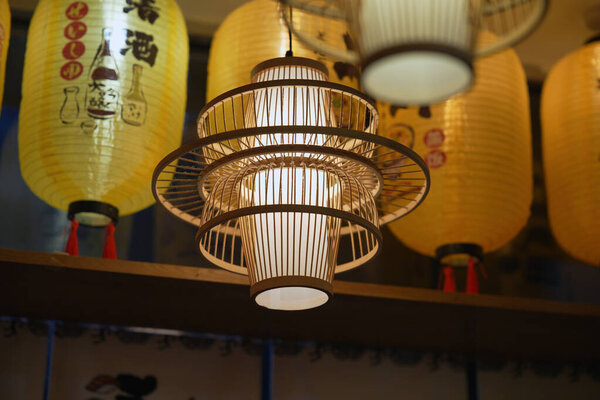 Мягкие желтые фонари висят над головой, бросая теплое свечение. Ниже, уникальный деревянный светильник придает очарование уютной атмосфере японского ресторана в вечернее время.