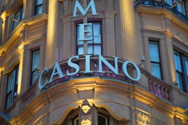 Bright lights illuminate the ornate facade of a historic casino building during the evening. The architectural details are enhanced by the warm glow from the lights.