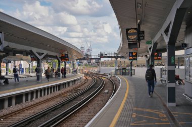 People are gathered on the platform of a busy train station in the daytime. The surroundings show multiple tracks and electronic displays indicating train information.