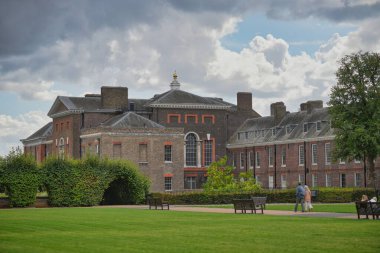 Visitors stroll through a vibrant green park, admiring a historic building in the background under a dramatic sky filled with clouds in London, England.