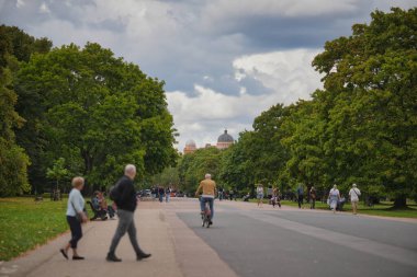 Visitors stroll and bike along a wide path in a green city park under a cloudy sky, with historic buildings peeking through the trees in the distance.