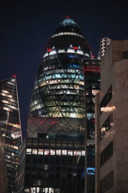 A striking modern skyscraper with a distinctive shape, illuminated against the night sky. The building stands among other contemporary structures in a vibrant urban setting.
