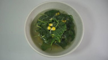 Hafif et suyu, yeşil yapraklı sebzeler ve dağınık sarı mısır çekirdekleri ile dolu bir kasenin tepe üstü görüntüsü..