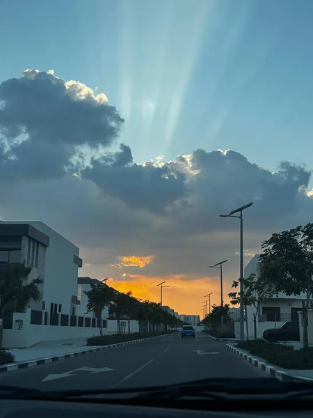 Dramatik günbatımı ışınları bulutları delip geçer. Modern binalar ve palmiye ağaçları ile kaplı bir sokağı aydınlatır..