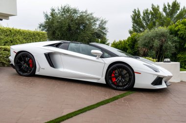 Beyaz Lamborghini Aventador Roadster bir garaj yoluna park etmiş, parlak tasarımını ve kırmızı fren kalibrelerini gösteriyor..