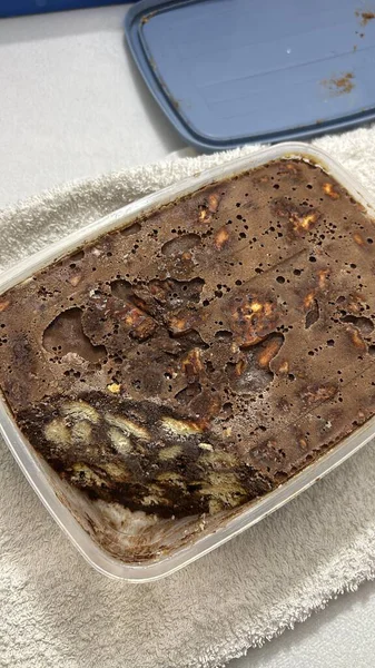 Dikdörtgen plastik bir kapta büyük, kısmen yenmiş çikolatalı bir tatlı ve görünür bisküvi parçaları bulunur. Tatlı yoğun ve zengin görünüyor..