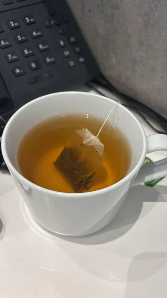 İçi demlenmiş çay dolu beyaz bir kupa ve içinde hala çay poşeti var. Telefonun bir kısmı arka planda görünür..