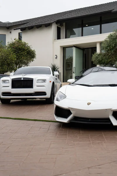İki lüks beyaz araba, bir Rolls Royce ve bir Lamborghini, modern bir evin önüne park edilmiş..