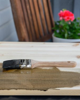 Tahta saplı bir boya fırçası, koyu ahşap bir leke sürmenin ortasında, açık tahta bir yüzeyde bulunur. Arkaplanda tahta bir sandalye ve saksıda kırmızı bir çiçek bulanıklaşıyor..