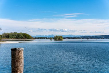 Bir rıhtımdan Constance Gölü 'ne bakın. Sahile uzanan çakıl taşlarını gösteriyor. Arka planda kar kaplı Alpler ve mavi gökyüzü var..