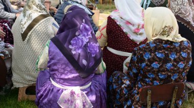 Alevi kadınlarının canlı çiçek desenli başörtüleri ve desenli elbiseleriyle dış kültür toplantılarında düşük sandalyelerde otururken Anadolu mirası ve toplum tahvillerini ön plana çıkardıkları arka plan.