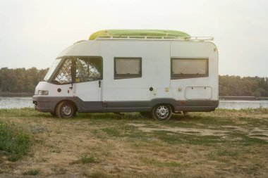 Çimenli Tuna nehri kıyısında park etmiş beyaz karavanın yan görüntüsü, sıcak akşam ışığı altında parlak yeşil kanoyla dolu çatı, açık havada seyahat özgürlüğünü ve vanhayat macerasını simgeliyor..