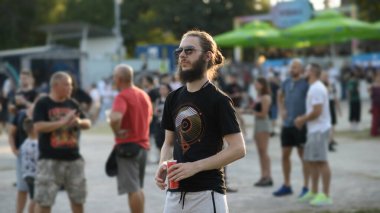06 / 25 / 2025 - Sofya, Bulgaristan: Güneş gözlüğü ve sakallı adam açık hava etkinliğinde içki kutusu tutuyor. Siyah bir grafik gömlek ve şort giyiyor. Bulanık arkaplanda insanlar görünür.