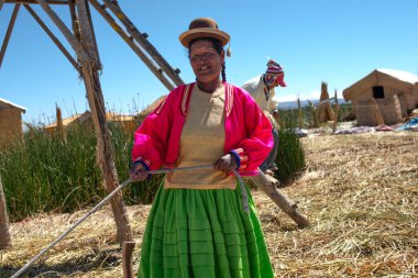 Uros Adaları, Peru - 10 / 16 / 2012: Peru 'daki Titicaca Gölü' nün yüzen adalarında yaşayan yerli bir kadın bowling şapkası, pembe ceket ve yeşil etekle arka planda kamış kulübeleri olan bir ipi çekerken geleneksel giysiler giyiyor.