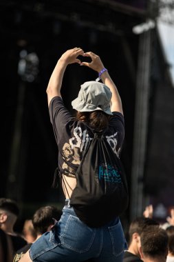 07 / 25 / 2025 - Plovdiv, Bulgaristan: Kova şapkalı kadın, siyah bantlı tişört, kot şortlu ve Hills of Rock sırt çantası dolu bir festival kalabalığında omuzlarına oturmuş, açık hava müzik konserinde sahneye kalp şeklinde bir el hareketi yapıyor.