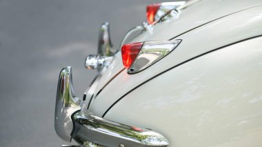 Eski bir fildişi arabanın arka çamurluğunda cilalanmış krom tampon ve kırmızı stop lambası klasik bir otomobil fuarı sırasında sessiz bir yolda ağaçları yansıtıyor..