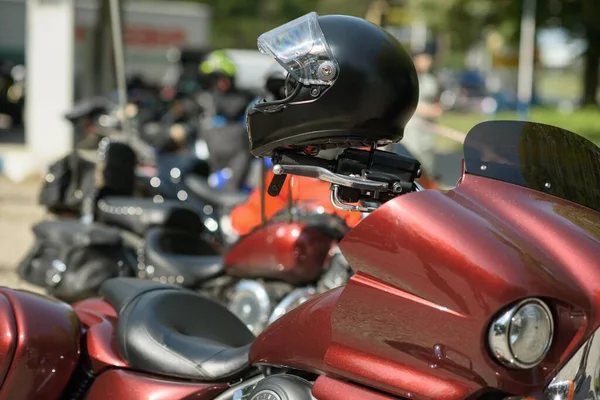 Odaklanmış siyah kasklı kırmızı motosiklet ve gündüz vakti dışarıda park etmiş bir grup motosiklet, araç detaylarını, binicilerin güvenlik ekipmanlarını ve motosiklet topluluğunun içeriğini ön plana çıkarıyor..