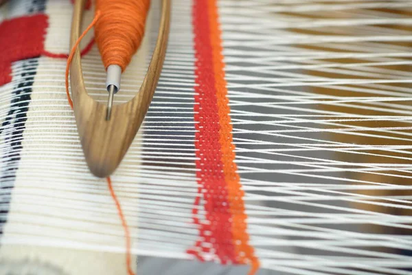 Parlak turuncu yünlerle dolu tahta mekik dar warp ipliklerinden geçer, el dokuma tezgahında canlı bir çizgi oluşturur ve geleneksel zanaatkar dokumasını gösterir.