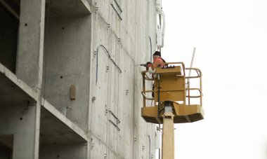 03 / 28 / 2025 - Bulgaristan 'ın Ruse kenti: Bir inşaat işçisi, çok katlı bir binanın beton duvarını delerken sarı bir asansörün üzerinde duruyor. Görüntü yüksek rakımlı teknik çalışmaları ve dikey site ekipmanları kullanımını gösterir.