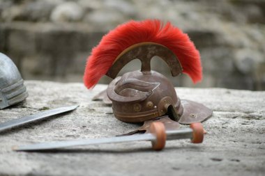 Üzerinde parlak kırmızı bir arma bulunan bronz bir Roma miğferi, yakınında kılıçlar bulunan taş bir yüzeyde duruyor. Bu da antik savaş ve tarihi canlandırmaların ruhunu çağrıştırıyor.