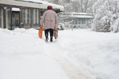 Rusçuk, Bulgaristan - 02 / 18 / 2025: Bir alışveriş çantası ve bir somun ekmek taşıyarak temiz ve karlı bir yolda yürüyen yaşlı bir kişi. Kışlık palto, bot ve kırmızı şapka giymiş bir adam, karla kaplı bir kafenin önünden geçiyor..