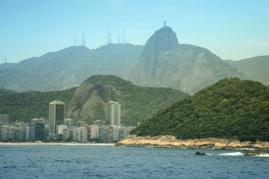 Brezilya 'nın Rio de Janeiro şehrinin manzarası, Corcovado Dağı' nın tepesindeki kurtarıcı heykeli ikonik İsa 'nın görüntüsü. Ön planda kıyı şeridi boyunca uzanan kentsel binalar, yemyeşil tepeler ve engebeli dağlar çarpıcı bir zemin oluşturuyor..