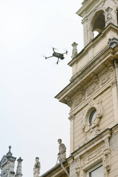 A modern drone flies near a Baroque-inspired historic building featuring intricate carvings and ornate statues, highlighting the contrast between advanced technology and classic architecture against an overcast sky.