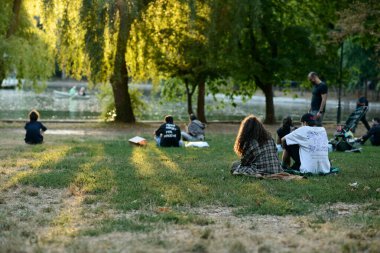 Bükreş, Romanya - 09 / 18 / 2025: Cismigiu Gardens, Bükreş, Romanya 'da göle bakan çimlerin üzerinde oturan gençler, yaz günü öğleden sonraları ağaçlar, güneş ışığı ve arka planda küçük bir tekne ile birlikte oturuyorlar.