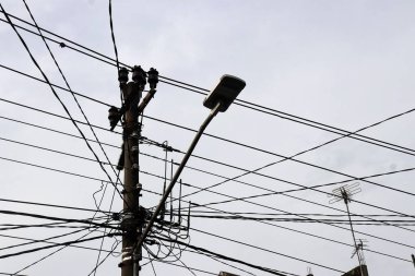 Temiz bir gökyüzüne karşı karmaşık kablolar ve paslı elektrik direkleriyle dolu karmaşık bir küme, kentsel elektrik altyapısının zorluklarını sembolize ediyor. Siyah kabloların yoğun ağı