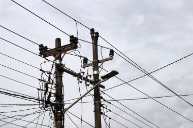 Temiz bir gökyüzüne karşı karmaşık kablolar ve paslı elektrik direkleriyle dolu karmaşık bir küme, kentsel elektrik altyapısının zorluklarını sembolize ediyor. Siyah kabloların yoğun ağı