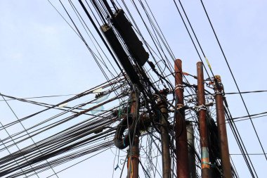 Temiz bir gökyüzüne karşı karmaşık kablolar ve paslı elektrik direkleriyle dolu karmaşık bir küme, kentsel elektrik altyapısının zorluklarını sembolize ediyor. Siyah kabloların yoğun ağı